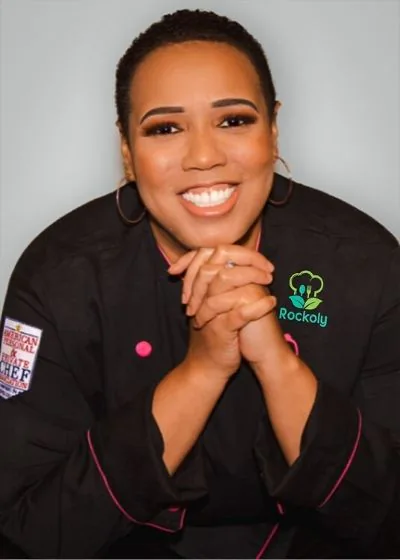 Chef A'Donna smiling with hands clasped in a pink-trimmed black chef coat with a competition patch