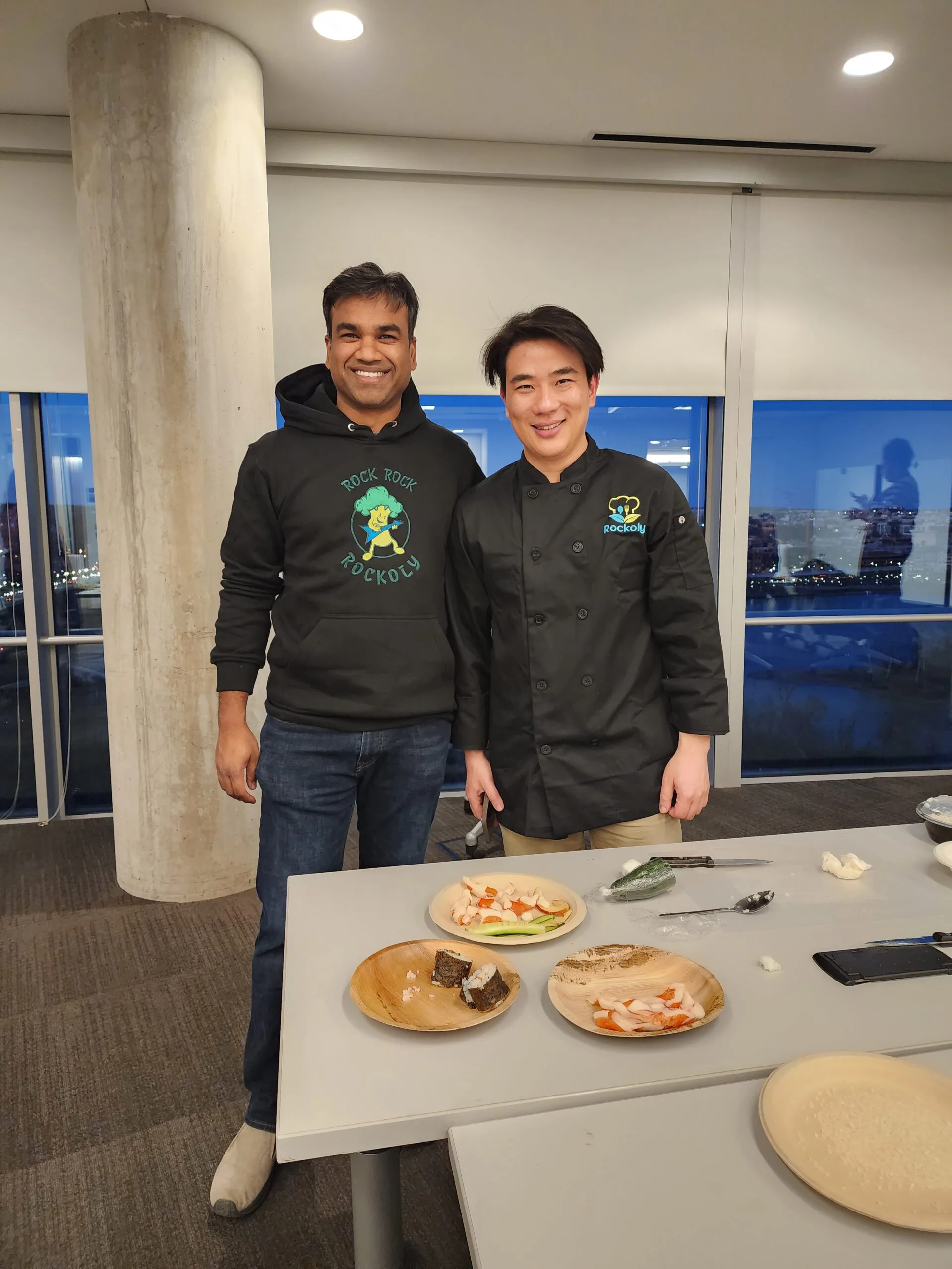 Chef and a guest standing beside wooden plates of the dishes they made, city windows behind them