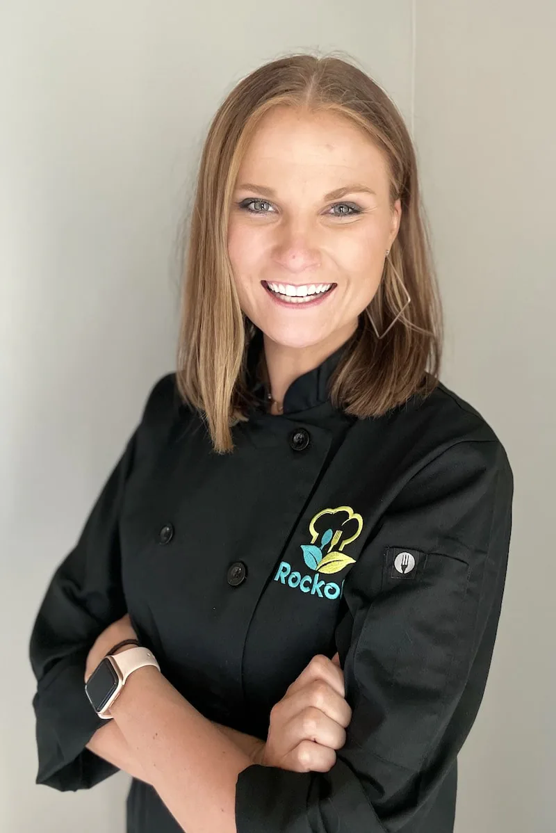 Chef Andrea smiling with arms crossed in a black chef coat embroidered with the Rockoly logo