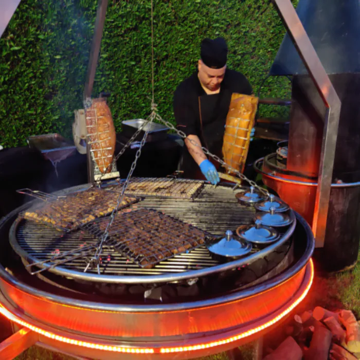 Chef tending meat on a glowing round wood-fired grill ringed with embers at an outdoor BBQ