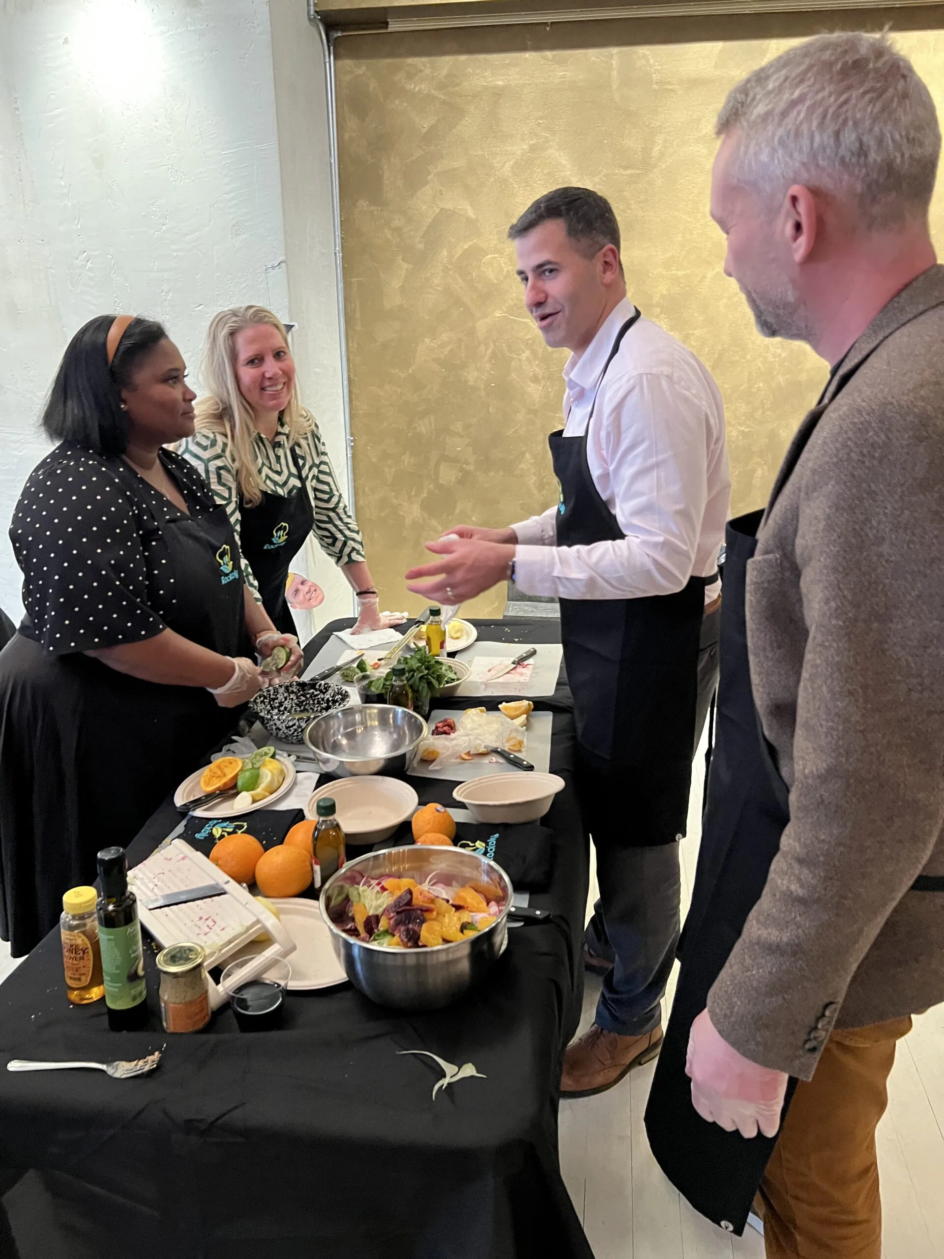 Chef in an apron talking with three guests beside a prep table of citrus, bowls and a mixing pot