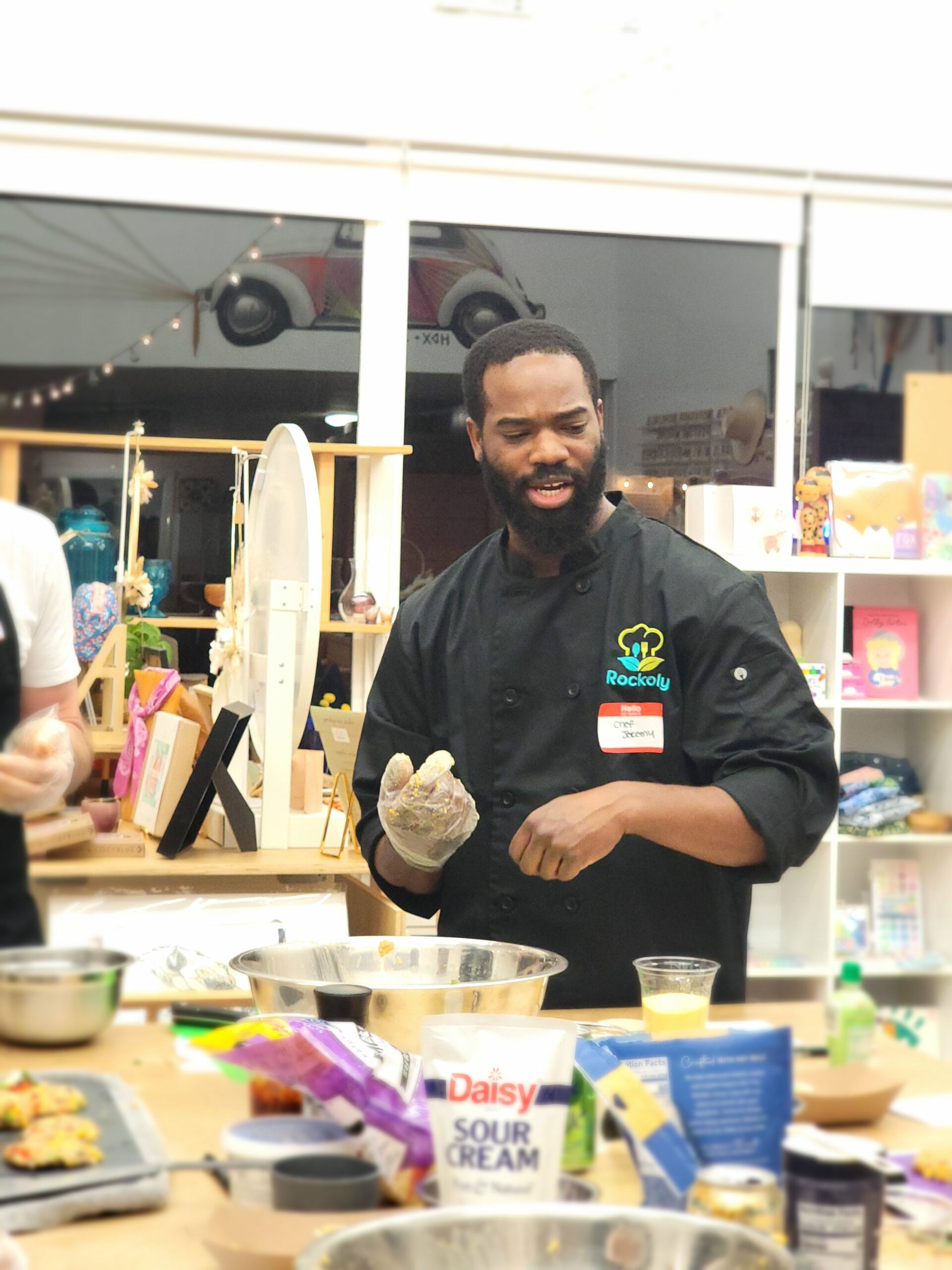 Bearded chef in a black Rockoly coat demonstrating a technique with gloved hands at an event