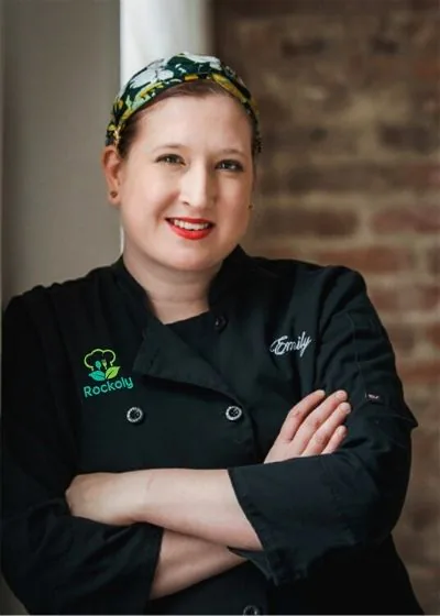 Chef Emily with arms crossed in a black chef coat and patterned headscarf beside a brick wall