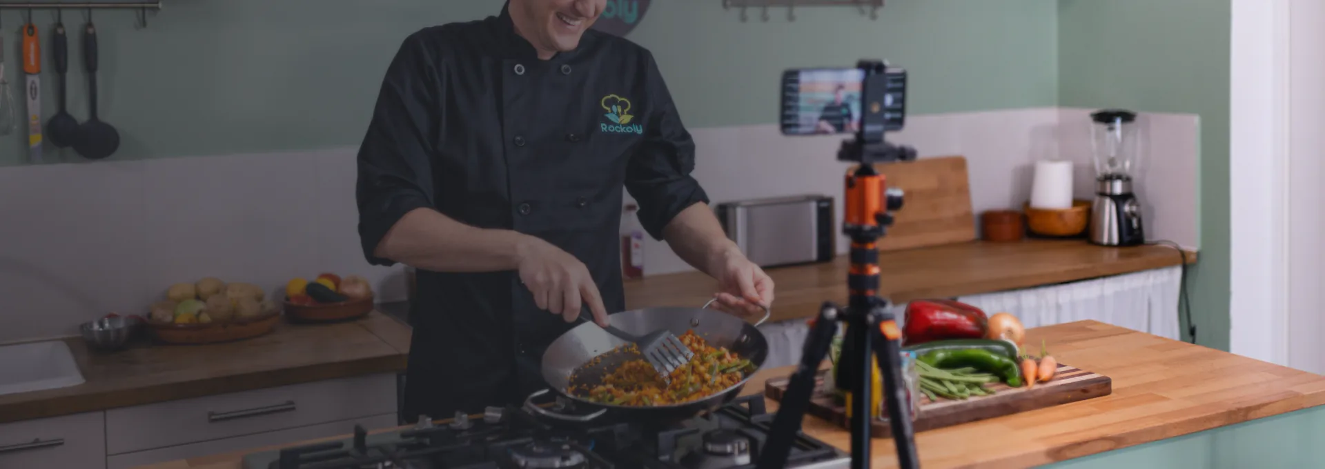 A chef cooks in a skillet while a phone on a tripod films him in a bright green kitchen