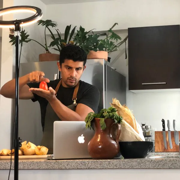 Chef Arturo holding a tomato in front of a ring light and laptop while filming a virtual cooking class