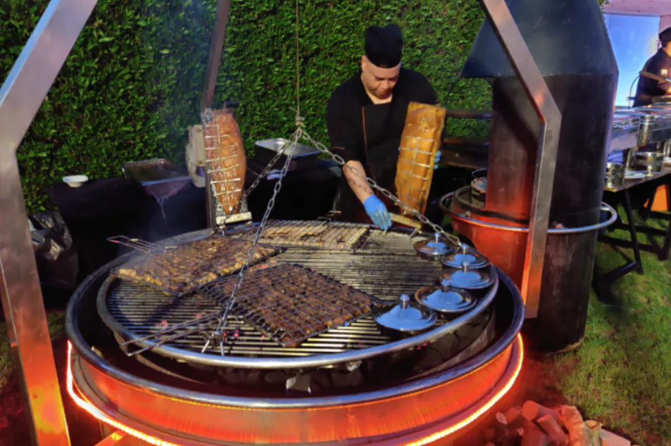 Chef grilling skewers and meat on a large round wood-fired barbecue at an outdoor event