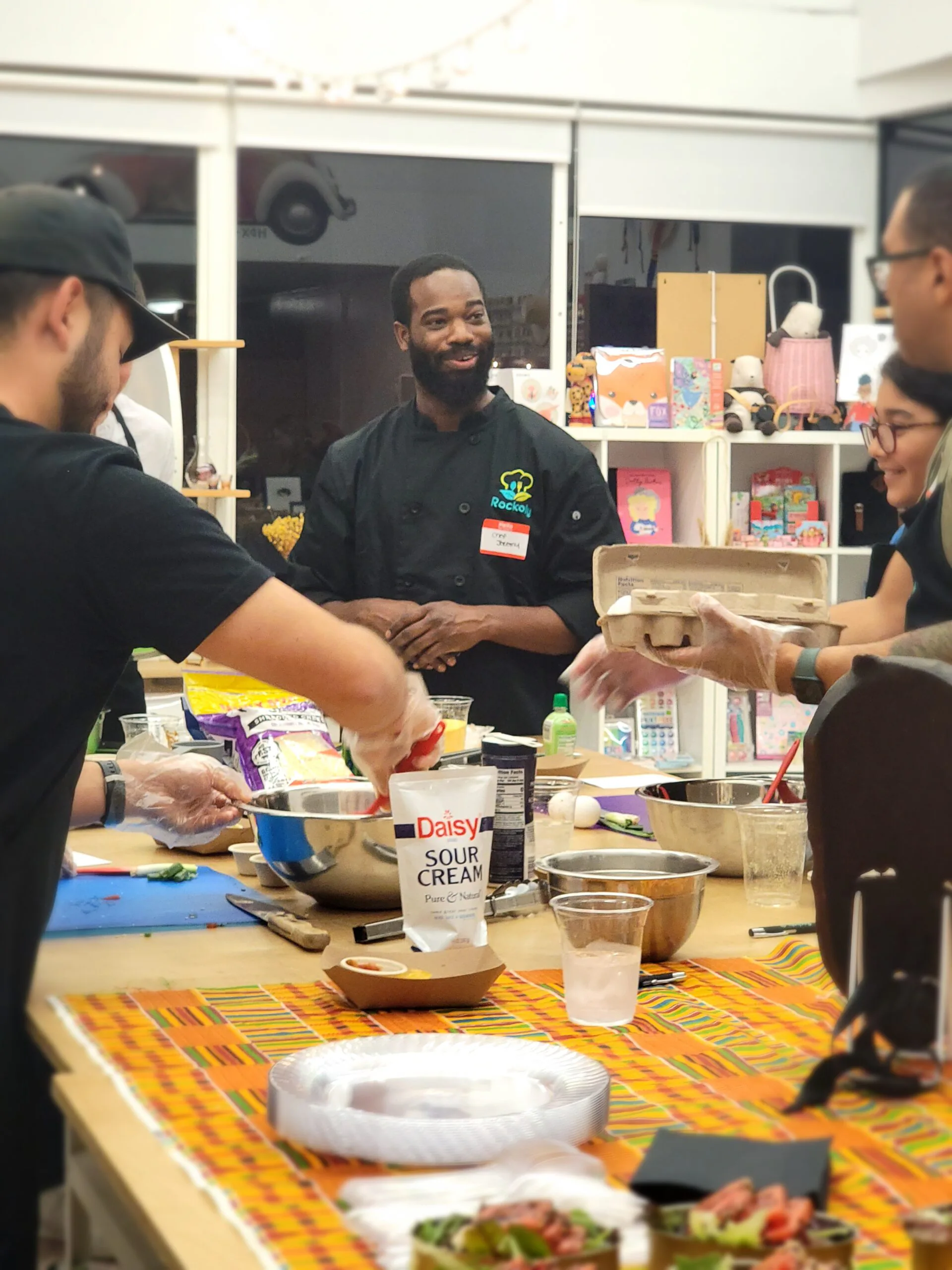 Chef in a black chef coat talking with guests around a prep table of bowls and ingredients