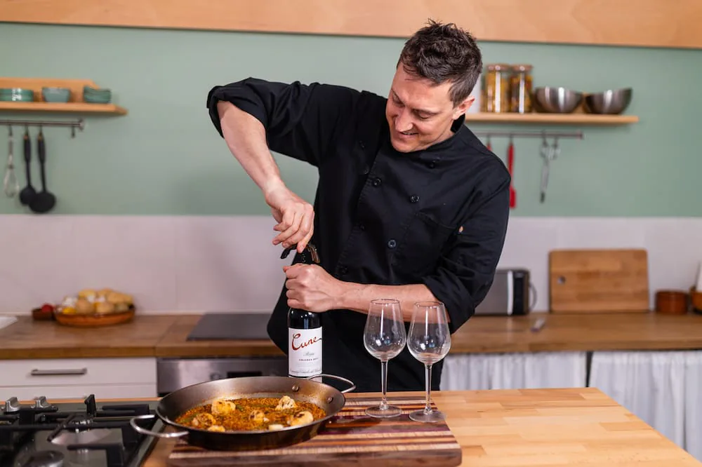 Chef Josh in a black chef's coat uncorking a bottle of wine beside a paella pan and two wine glasses