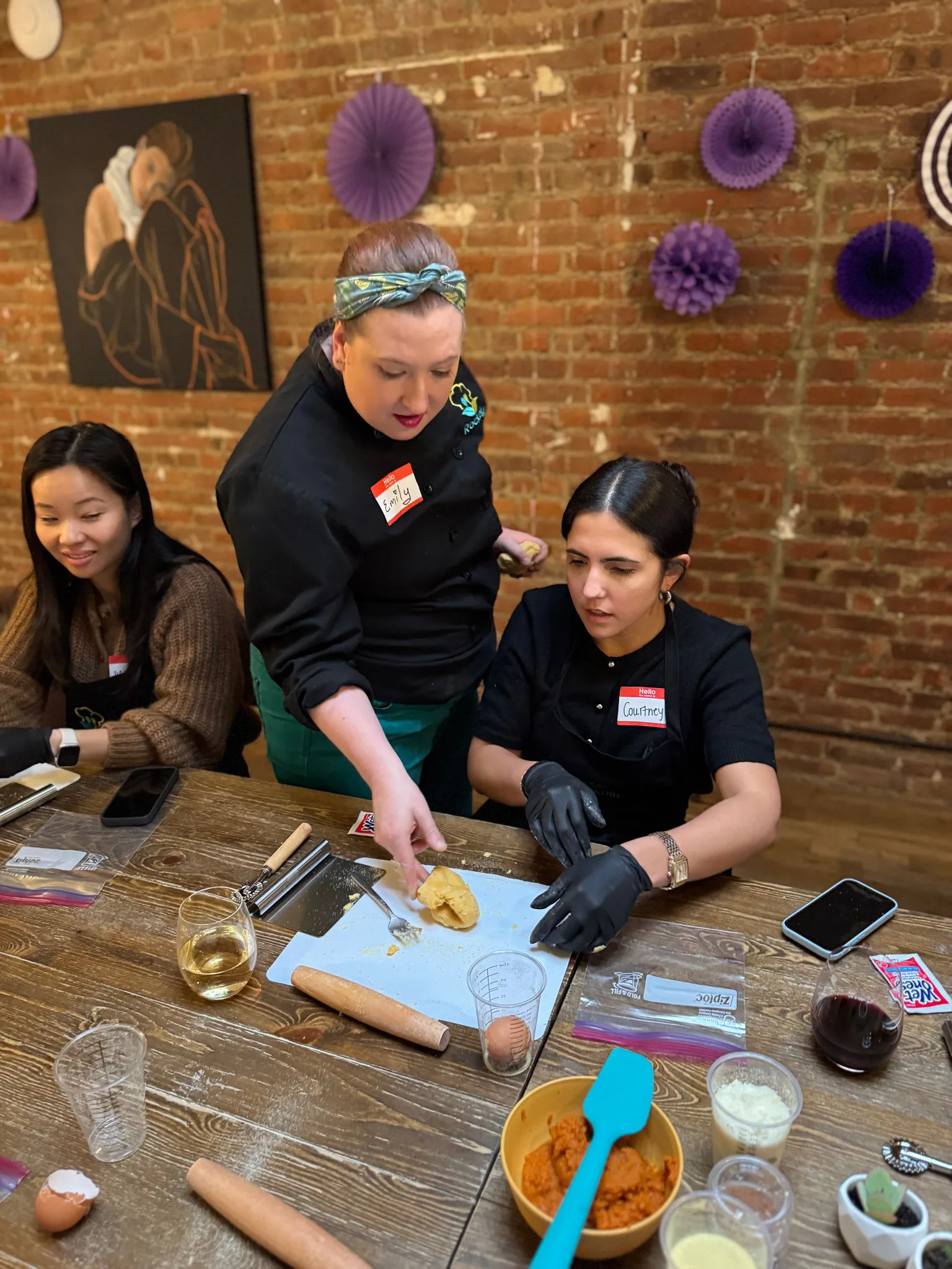 Chef in a green apron shaping dough at a table while two seated guests watch in a brick-walled studio