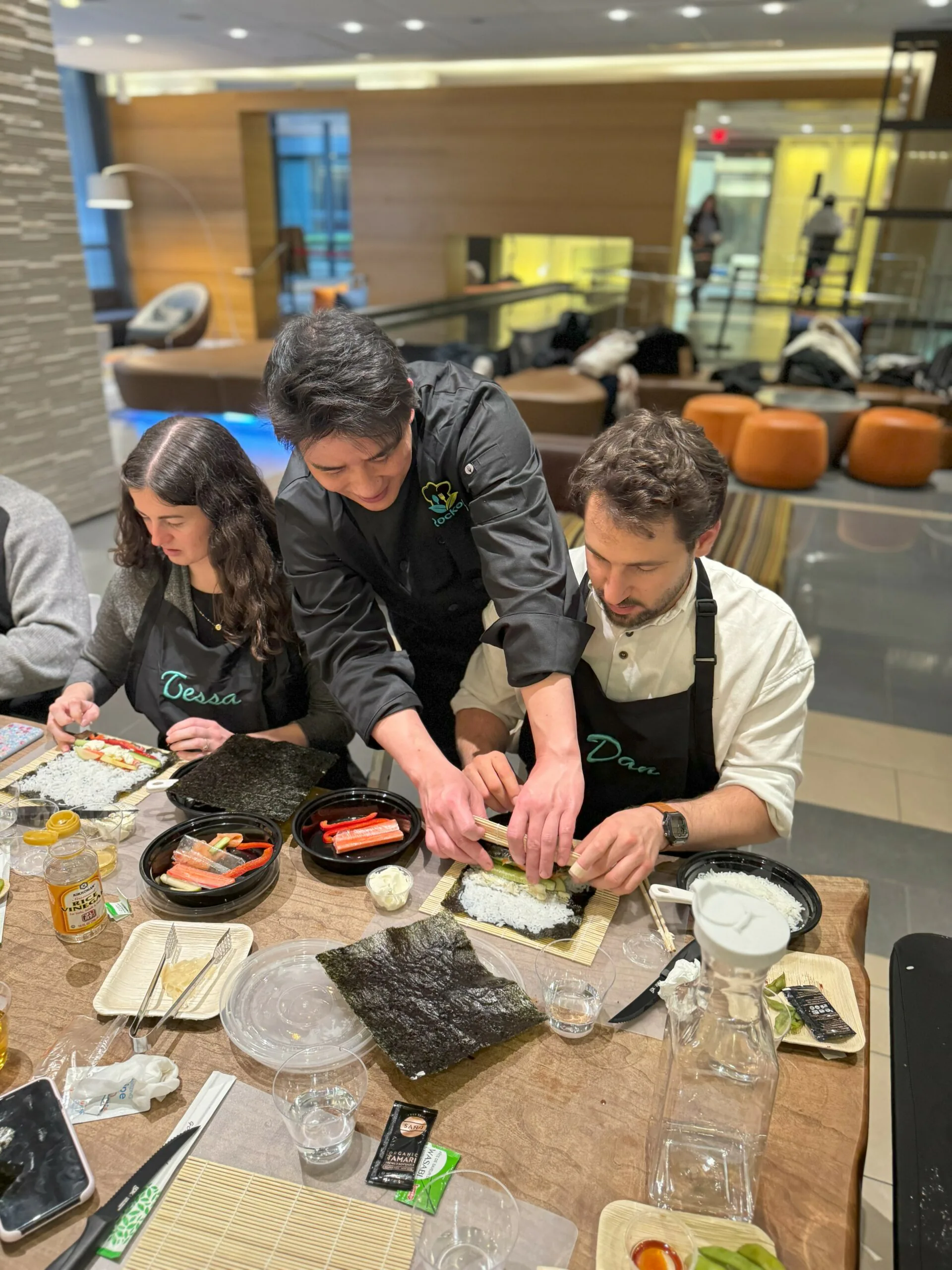 Chef leaning in to help two guests in aprons roll sushi with nori and rice at a table