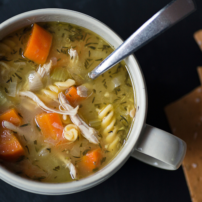 Mug of chicken noodle soup with shredded chicken, spiral pasta and carrots, crackers alongside