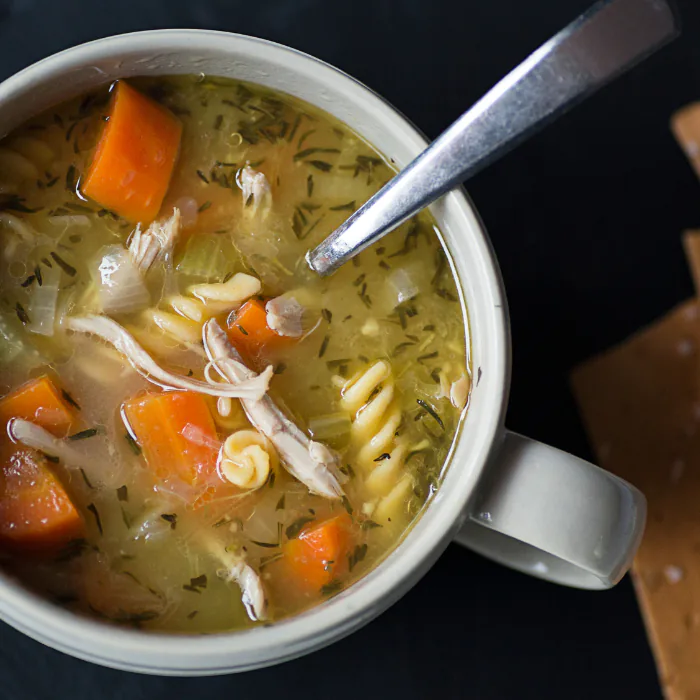 Mug of chicken noodle soup with shredded chicken, spiral pasta and carrots, crackers alongside