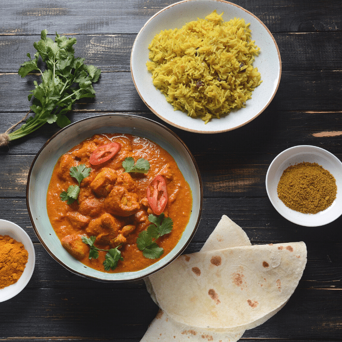 Bowl of chicken tikka masala with yellow rice, soft naan and small dishes of ground spices