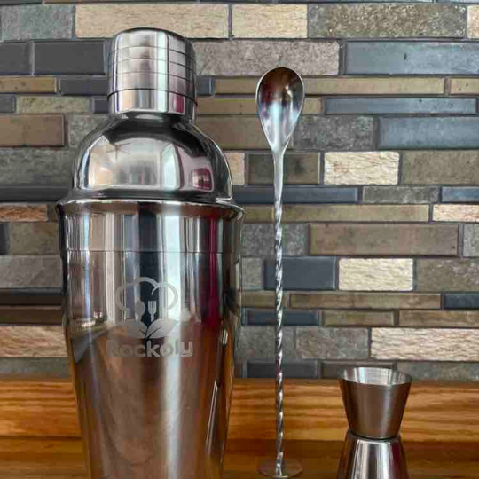 Stainless steel cocktail shaker engraved with the Rockoly logo, next to a twisted bar spoon and jigger
