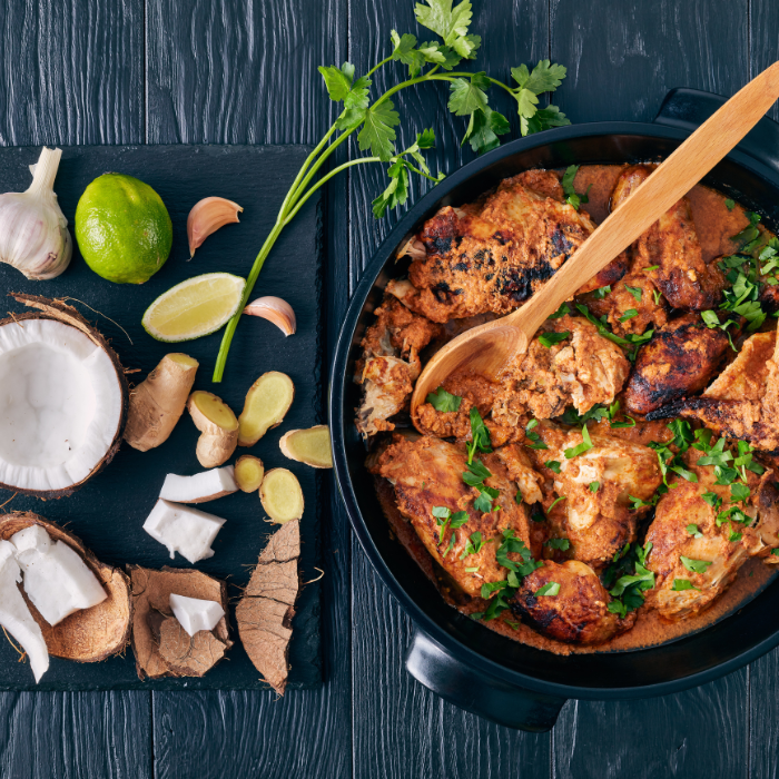 Pot of coconut chicken curry topped with cilantro beside lime, garlic, chilies and fresh coconut