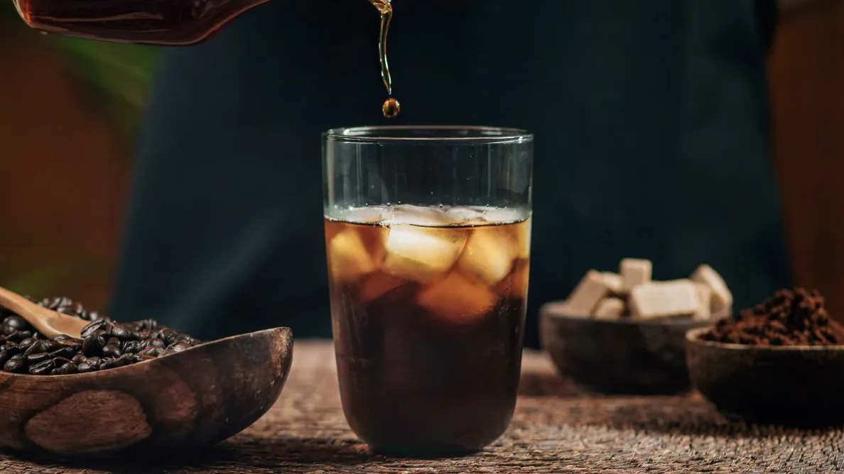 Cold brew coffee poured over ice into a tall glass, with coffee beans and sugar cubes in bowls