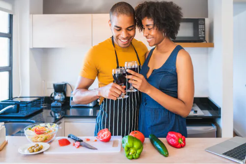 Couple clinking glasses of red wine in a kitchen with chopped peppers and tomatoes on the counter