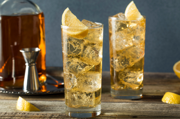 Two tall amber highballs over ice with lemon wedges, beside a whisky decanter and a jigger