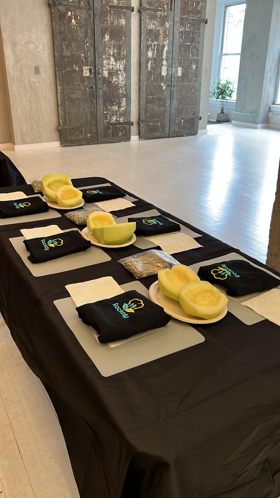 Long table set with folded aprons, cutting boards and halved yellow melons at each place setting