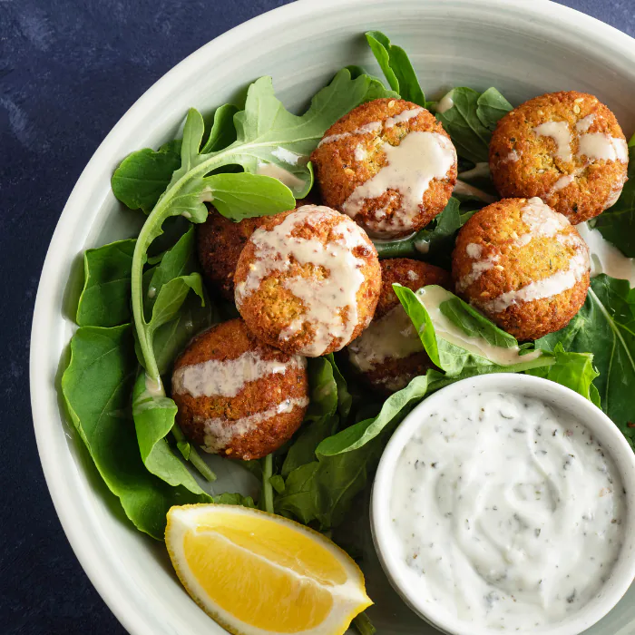 Crisp falafel balls on arugula drizzled with tahini, with a lemon wedge and a bowl of herbed dip