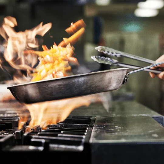 Flames leaping around a sauté pan of food being flambéed on a restaurant range