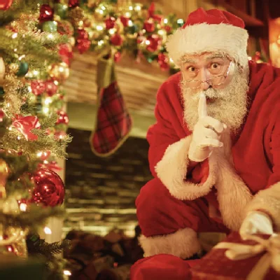 Santa Claus holding a finger to his lips beside a lit Christmas tree