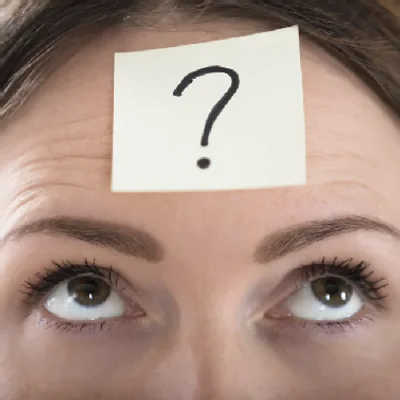 Close-up of a person's eyes looking up at a sticky note with a question mark stuck to their forehead
