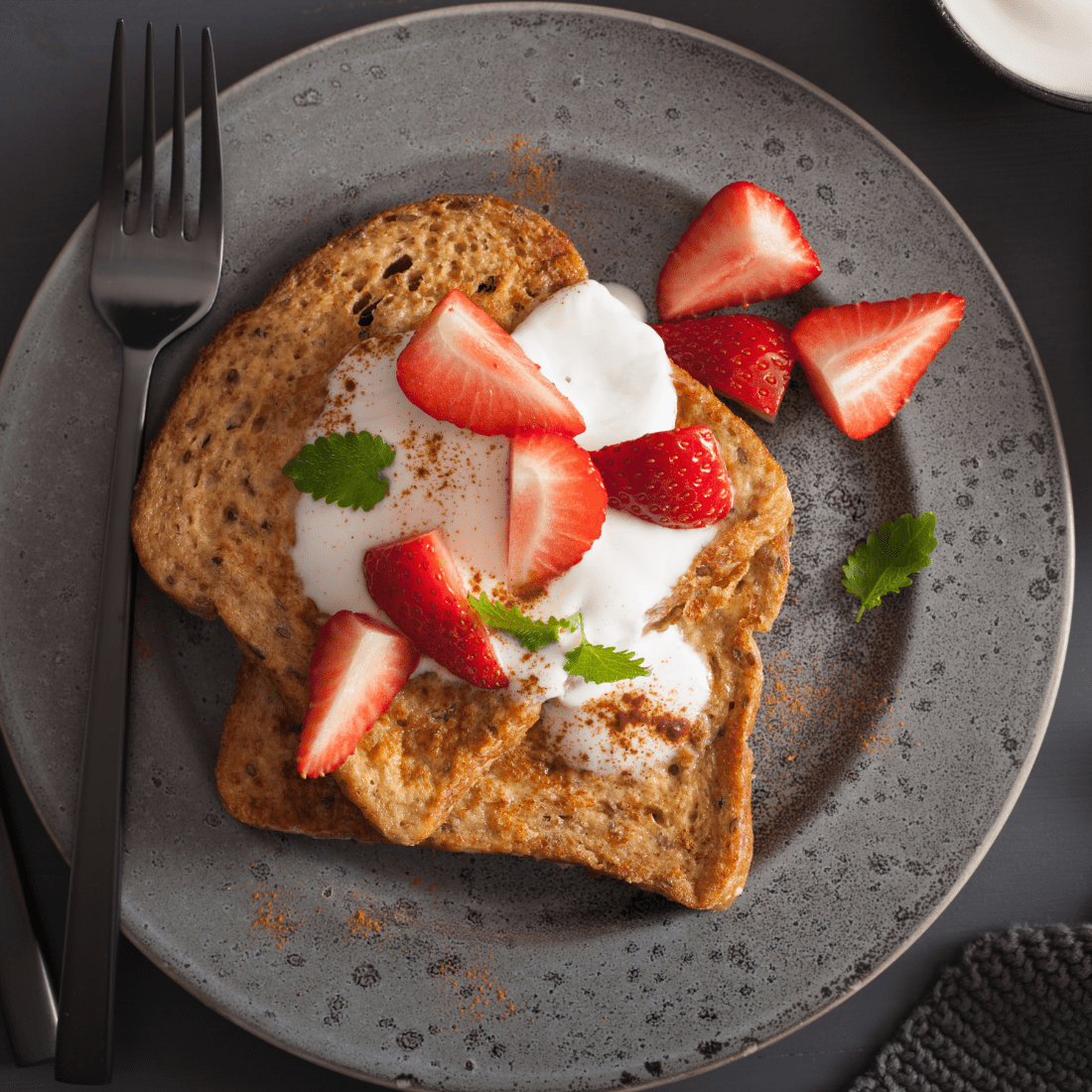 Slice of French toast topped with whipped cream, fresh strawberries, mint and cinnamon on a grey plate