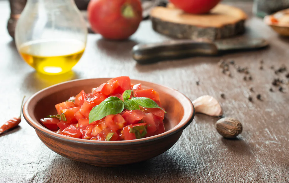 Rustic bowl of chopped tomato salsa topped with basil, with olive oil and garlic nearby