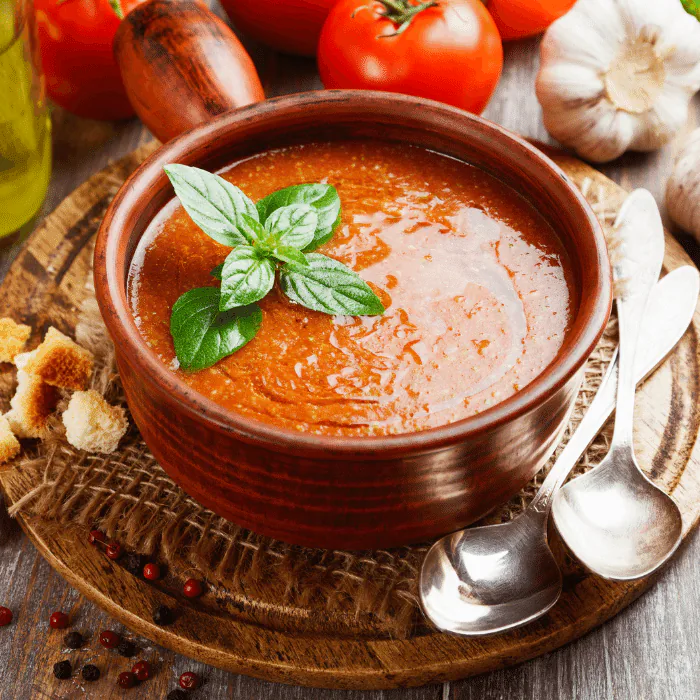 Bowl of chilled tomato gazpacho garnished with basil, with tomatoes, garlic and bread alongside