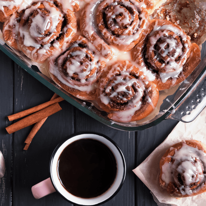 Tray of glazed cinnamon rolls beside cinnamon sticks and a mug of black coffee