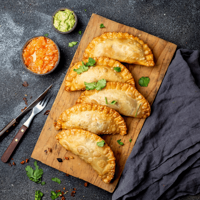 Row of golden crimped empanadas on a wooden board with bowls of red salsa and guacamole