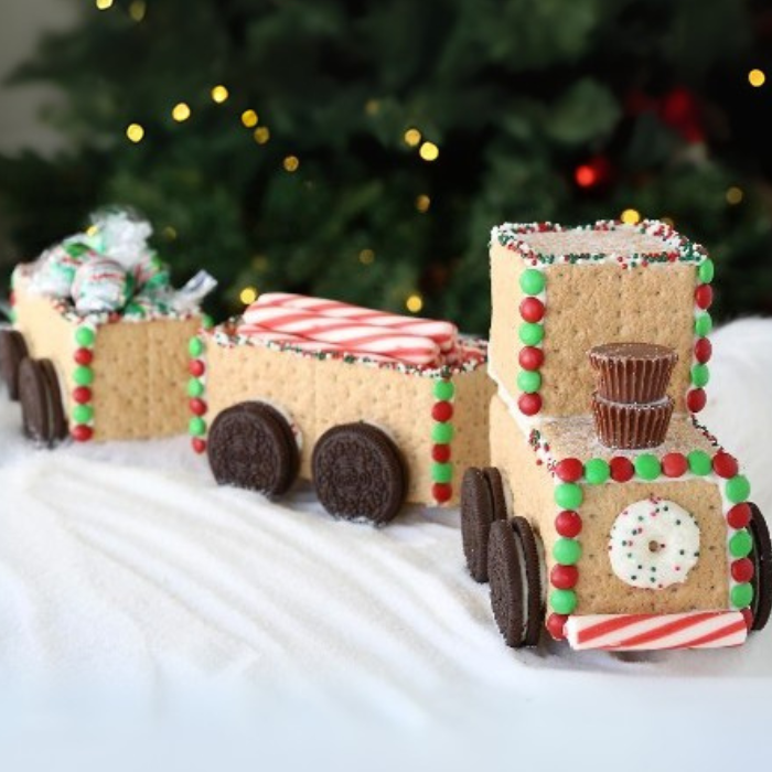 Graham cracker train decorated with candies and chocolate cookie wheels in front of a Christmas tree