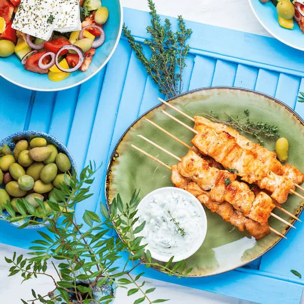 Grilled souvlaki skewers with tzatziki on a green plate, beside Greek salad and olives on a blue table