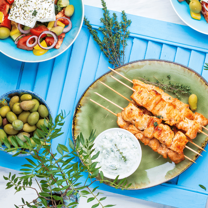 Grilled souvlaki skewers with tzatziki on a green plate, beside Greek salad and olives on a blue table