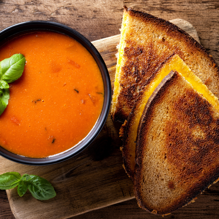 Grilled cheese sandwich halves with melting cheddar beside a bowl of tomato soup and basil