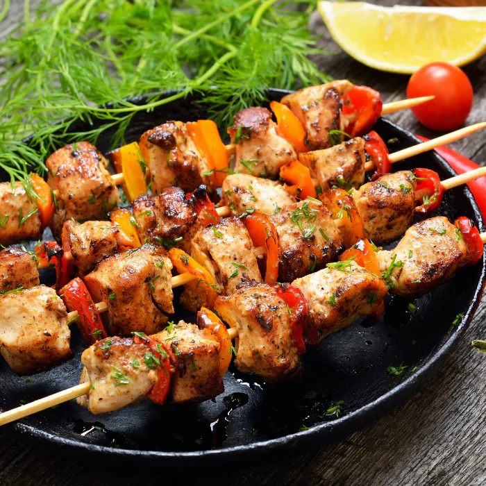 Grilled chicken and bell pepper skewers on a dark plate with dill, a lemon wedge and a cherry tomato