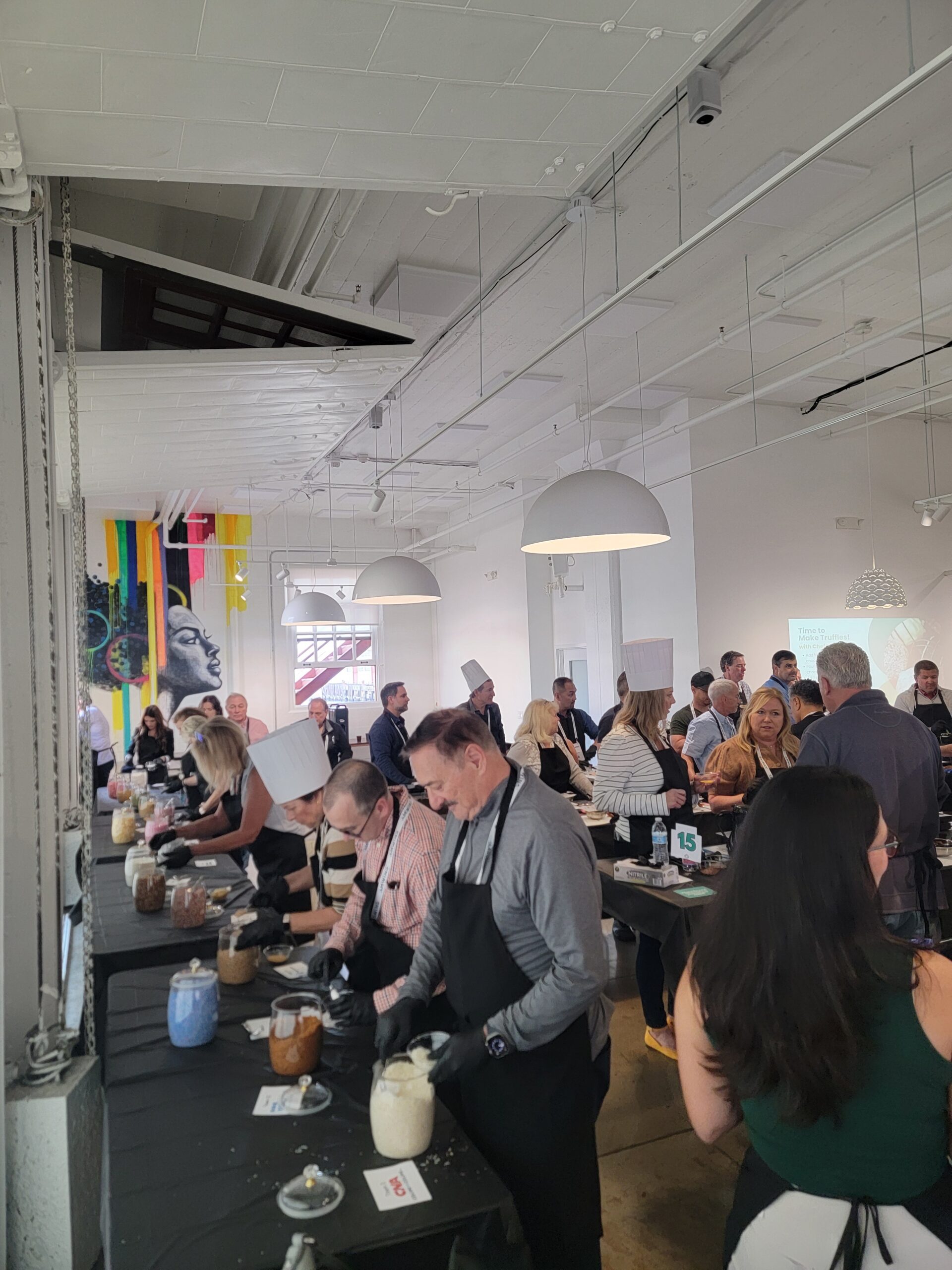 Guests in aprons working along rows of prep tables during a hands-on event in a bright loft