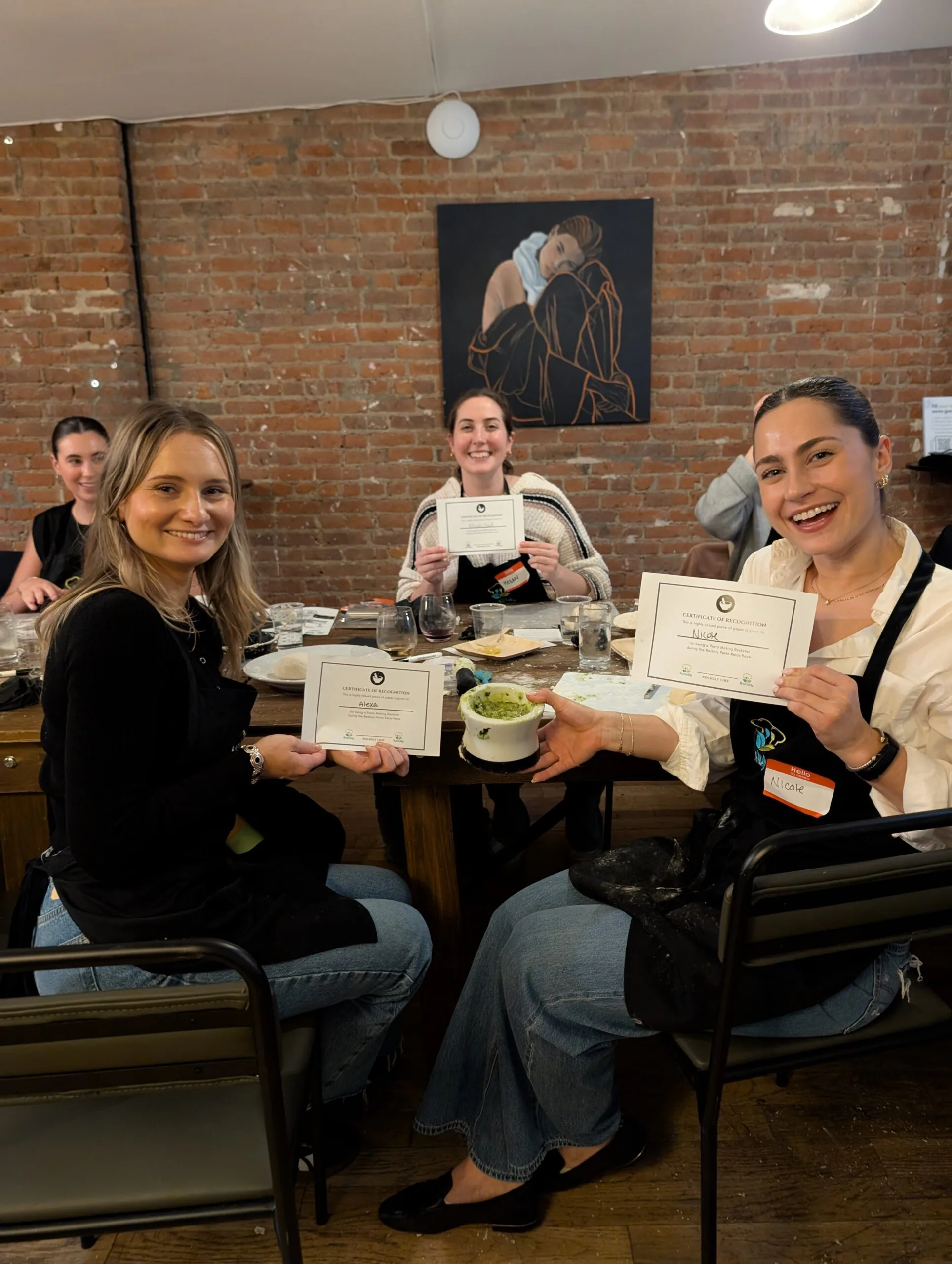 Three smiling guests hold printed certificates at a brick-walled table after a cooking event