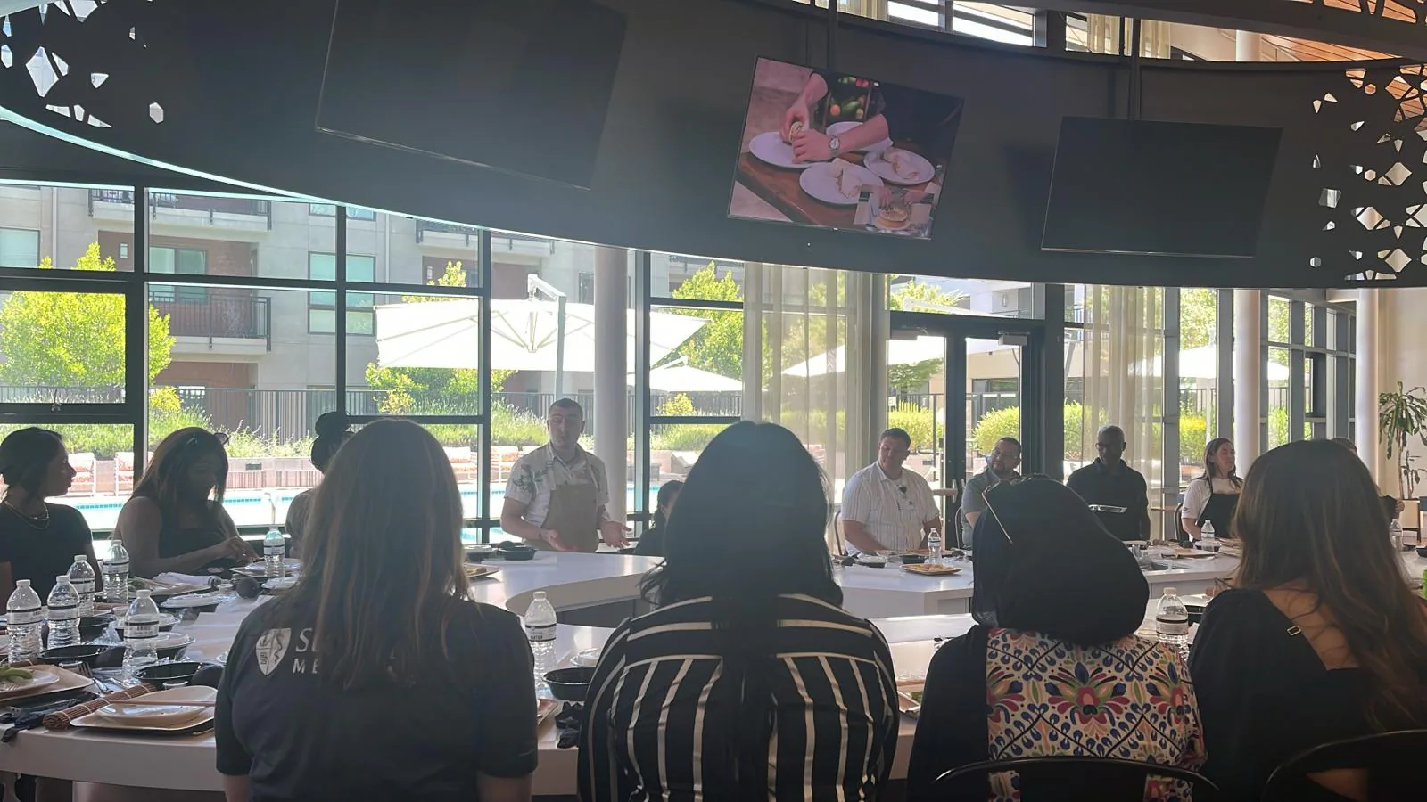 Guests seated around a table watch chefs demonstrate while a screen overhead shows the plated dish