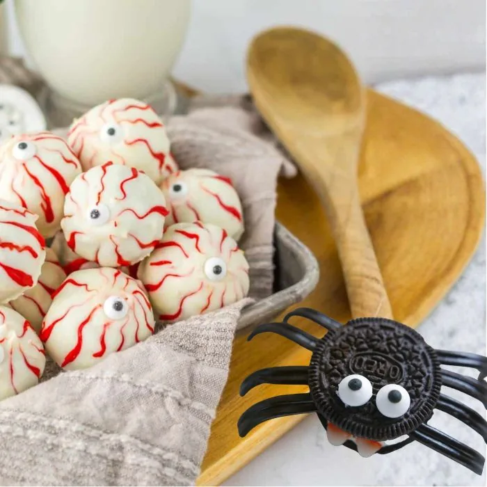 White chocolate eyeball truffles with red icing veins in a tin, beside an Oreo spider with candy-eye eyes on a wooden spoon