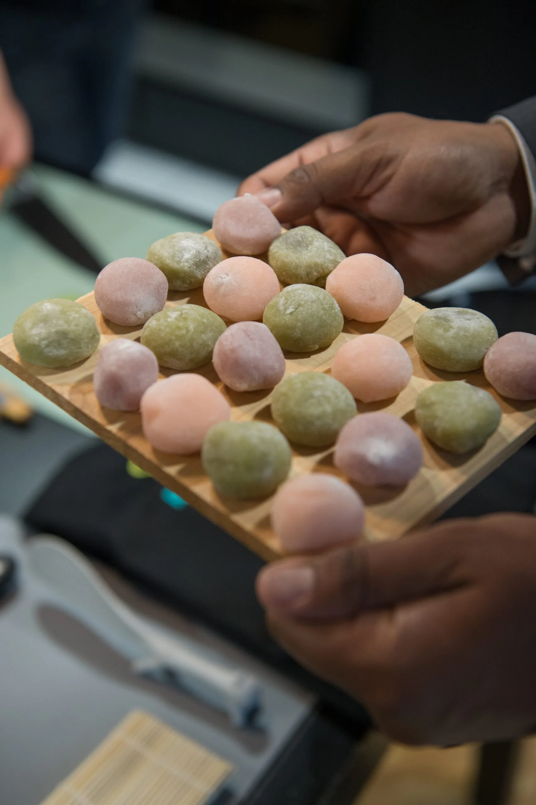 Hand holding a wooden board lined with rows of pink, green and purple mochi