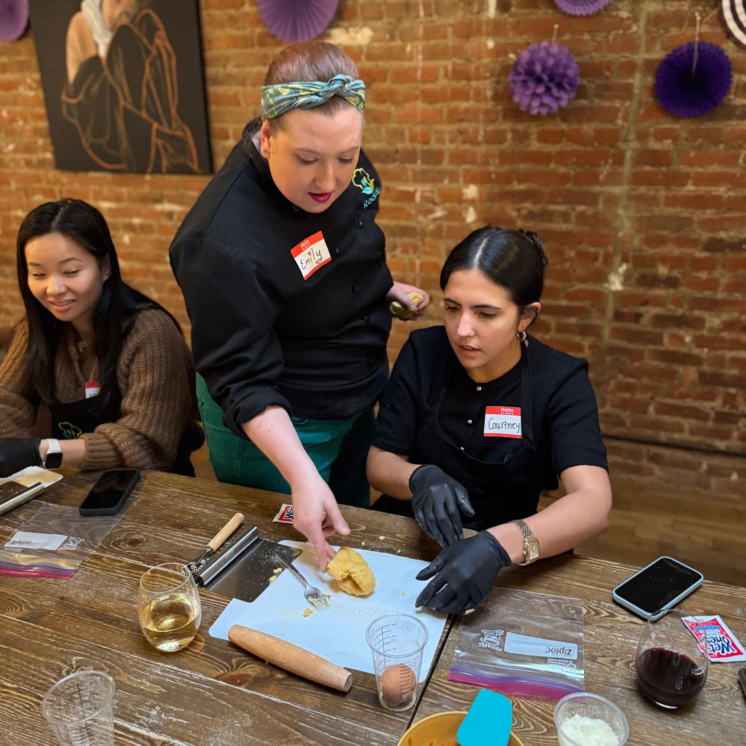 Participants at a hands-on cooking class rolling dough at a wooden table in a brick-walled event space