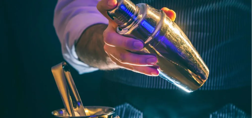 Hands shaking a gold cocktail shaker over a bar in low light