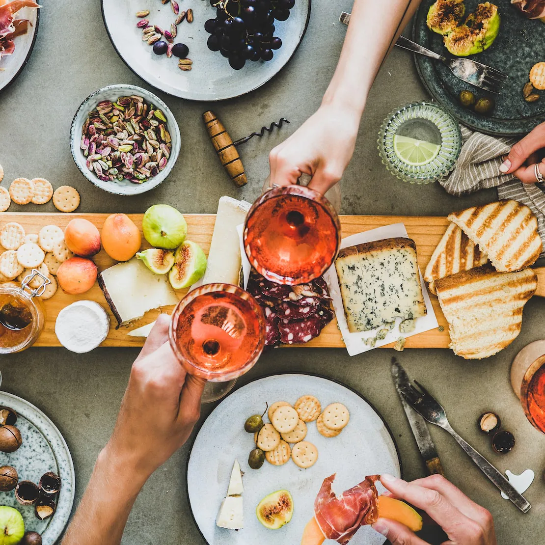 Two hands clinking glasses of rosé over a spread of cheese, grapes, figs, crackers and grilled bread