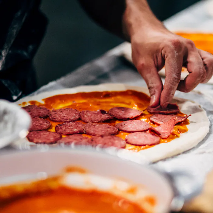 Hands arranging pepperoni slices over sauce on a raw pizza base dusted with flour