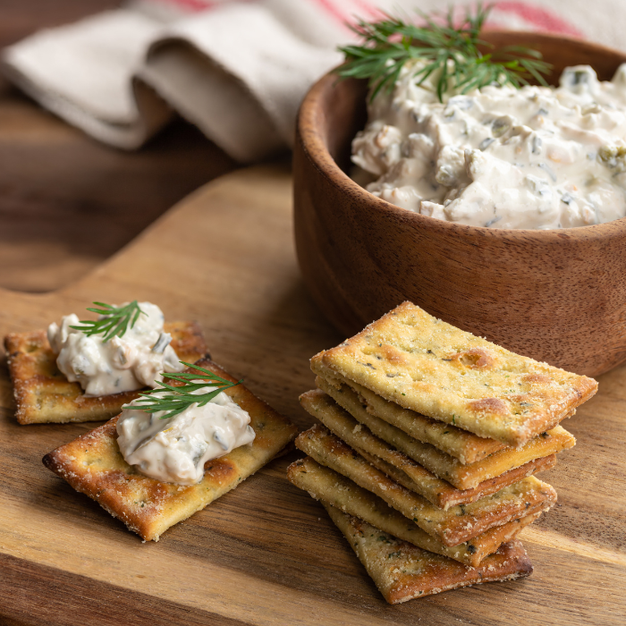 Homemade herb crackers on a wooden board with a bowl of creamy cheese dip topped with dill
