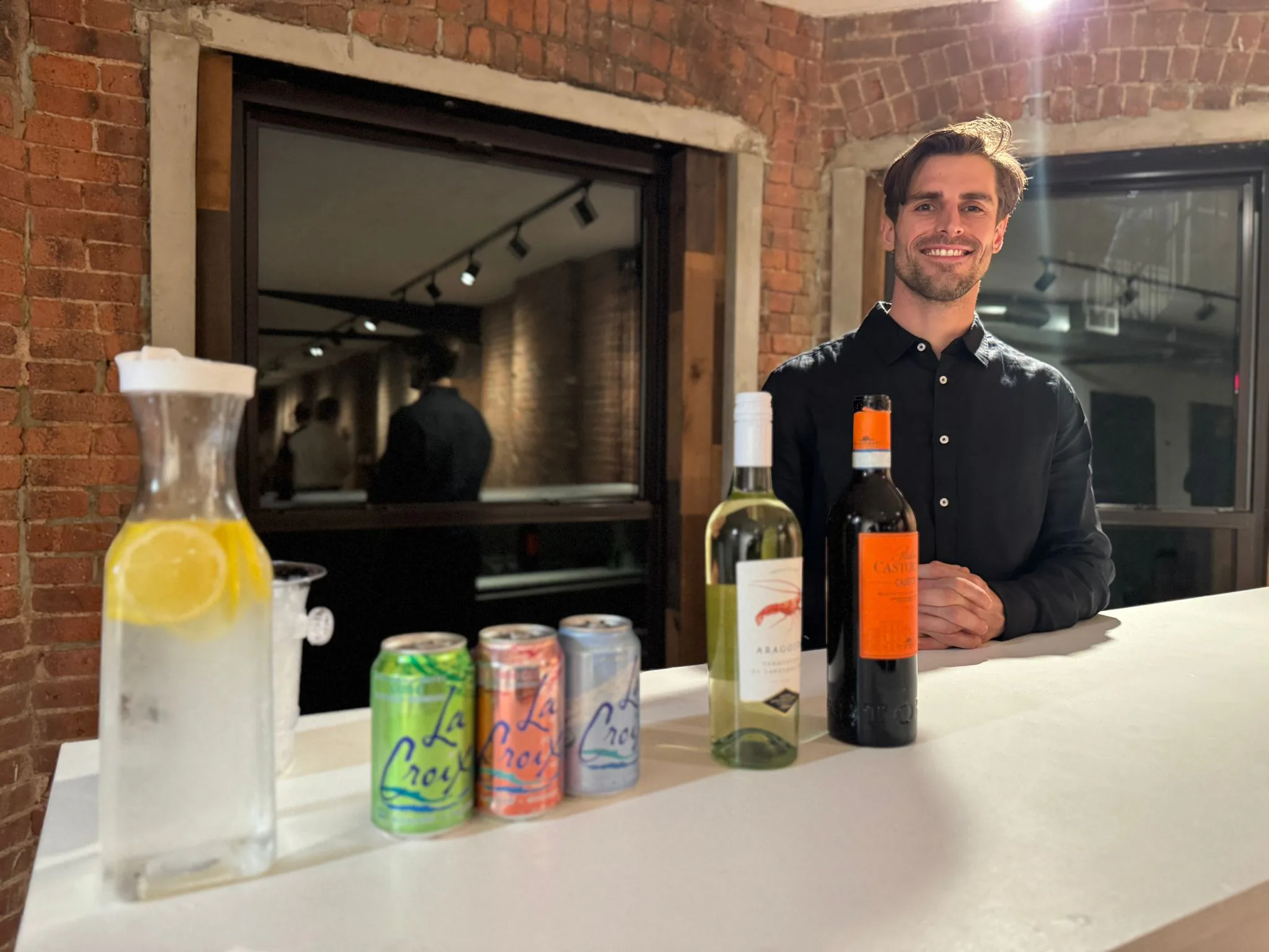 Smiling host behind a bar set with red and white wine, seltzer cans and a lemon water dispenser