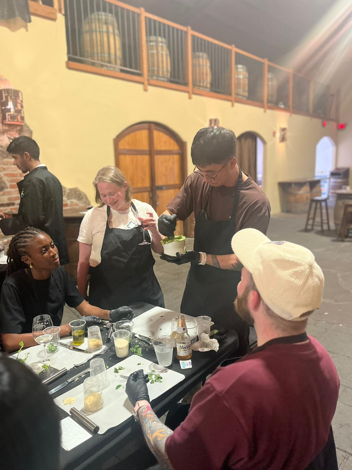 Guests in aprons gather at a prep table while a host plates greens at an in-person cooking event