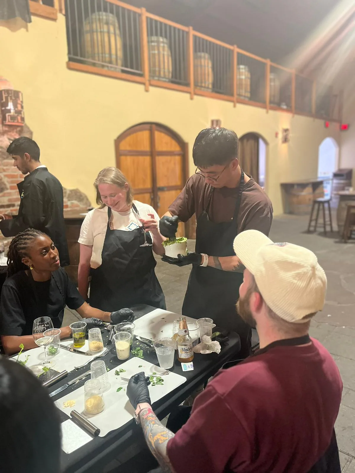 Guests in aprons gather at a prep table while a host plates greens at an in-person cooking event